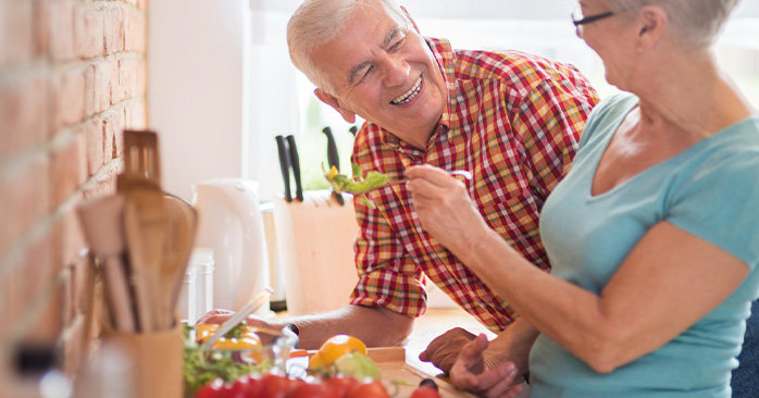 Elderly couple preparing vegetables in kitchen. Wife feeding husband lettuce.